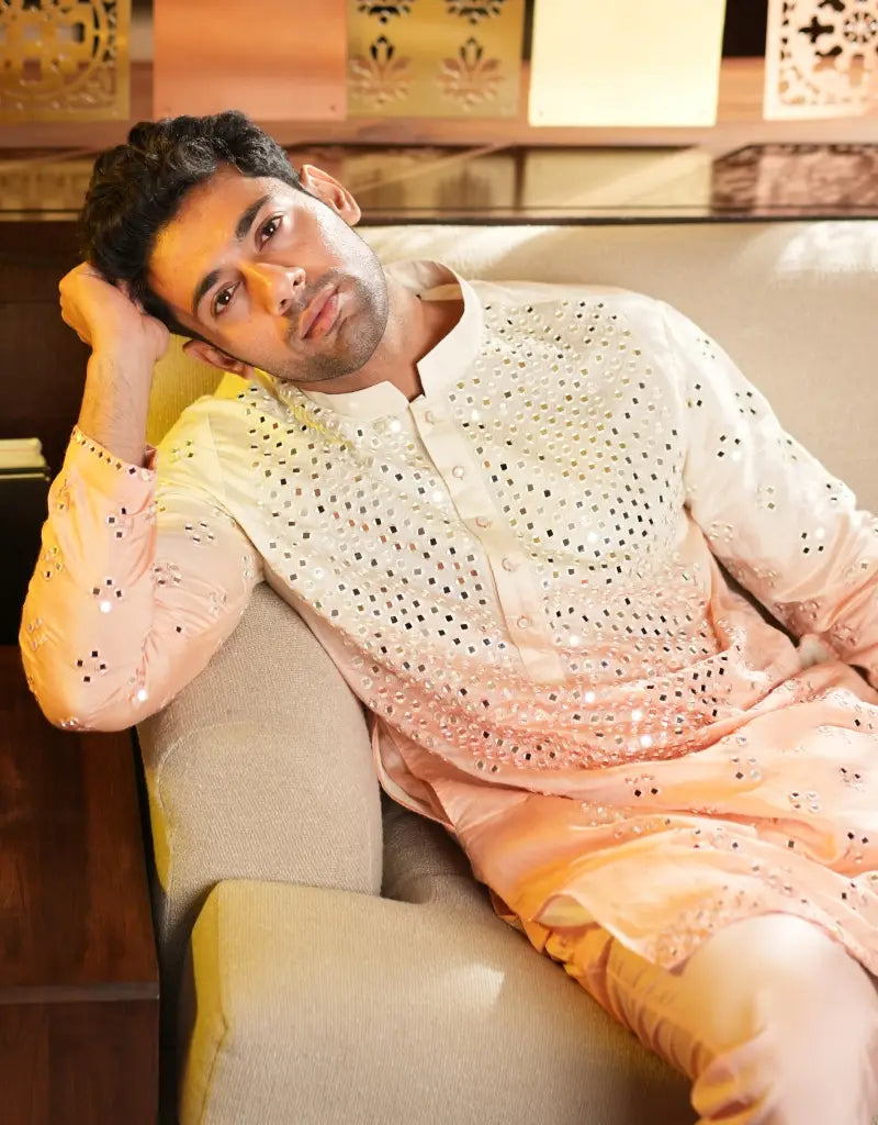A man wearing a light-colored traditional Blush Radiance Mirrorwork Kurta outfit sitting on a couch.