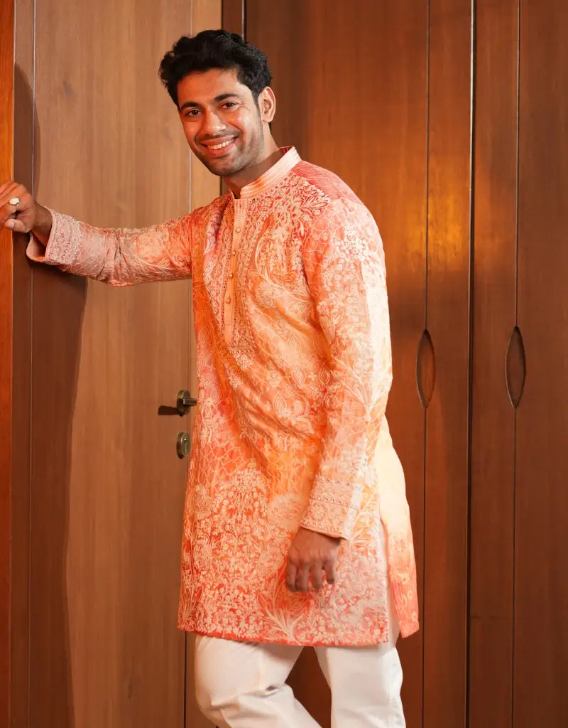 Man wearing an orange kurta with white pants standing against a wooden door.