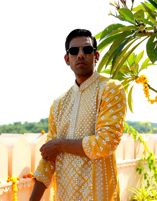 Man wearing a yellow and white patterned kurta with sunglasses, standing outdoors with greenery in the background.