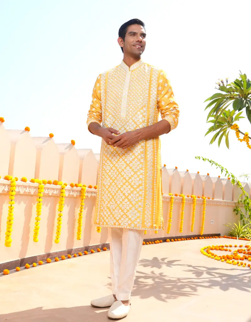 Man wearing a yellow kurta with white patterns standing outdoors with decorative elements.