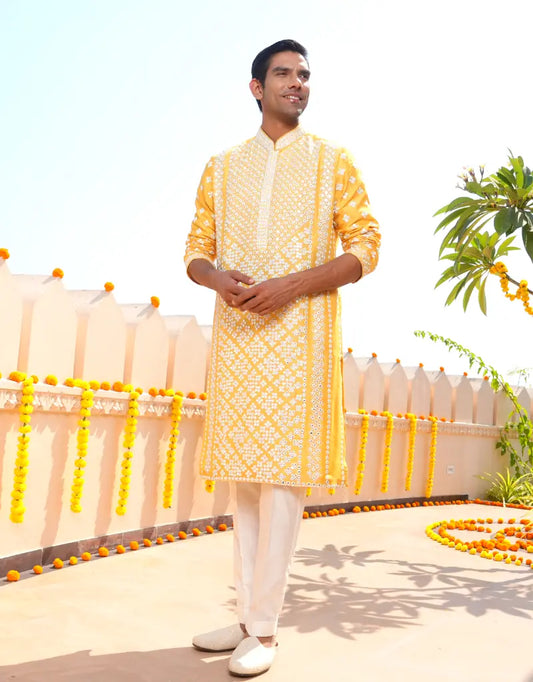 Man wearing a yellow kurta with white patterns standing outdoors with decorative elements.