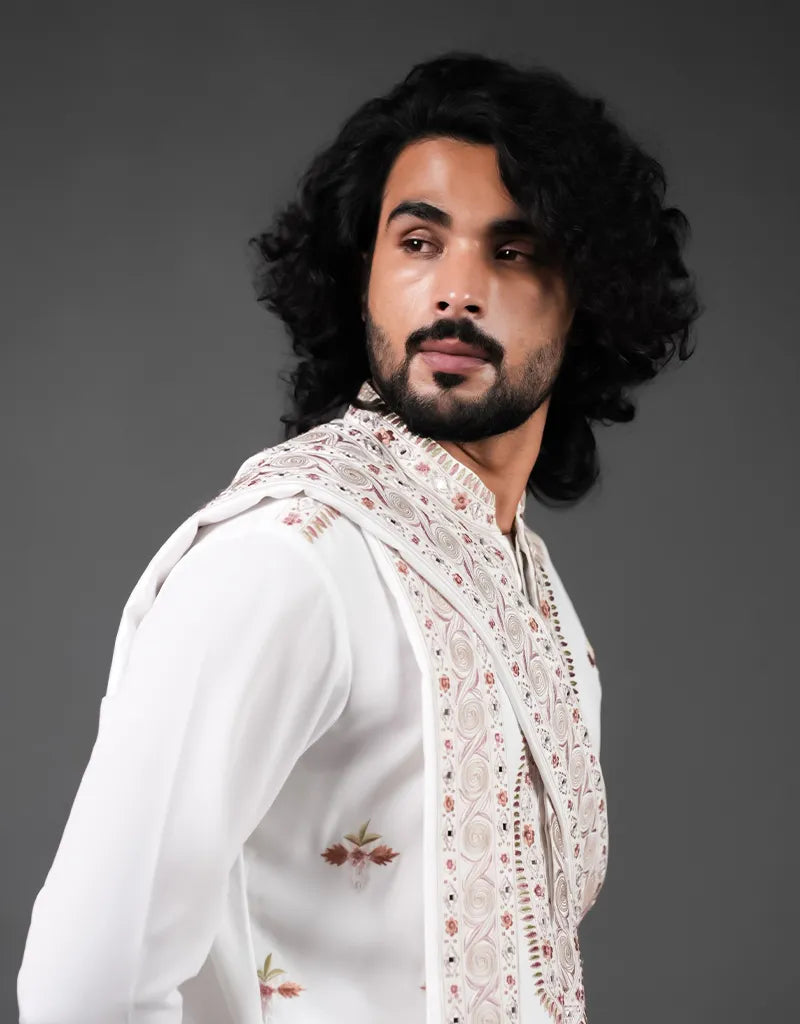 Man wearing a white traditional Kashmiri Mirror Kurta Set outfit with a patterned shawl against a dark background