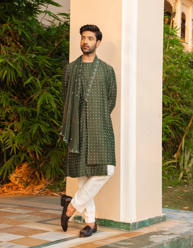 Man wearing a green kurta with white pants standing outdoors near a column.