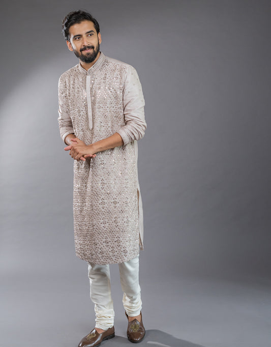 Man wearing a patterned one-piece satin silk kurta in light onion with light-colored pants against a gray background
