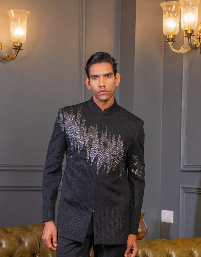 Man wearing a black embroidered Classic Bandhgala Indo-Western
in an elegant room with wall sconces.